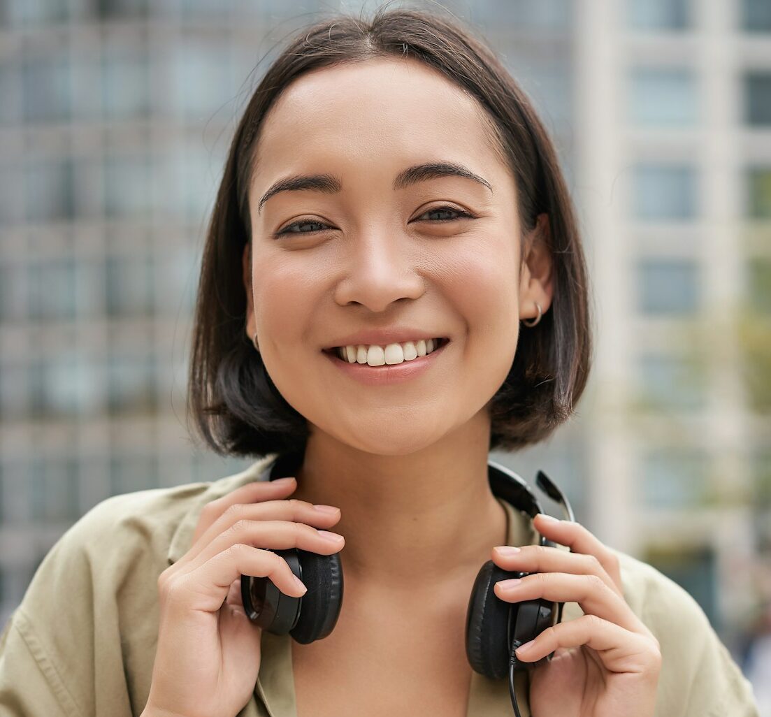 Portrait of smiling asian girl with headphones, posing in city centre, listening music